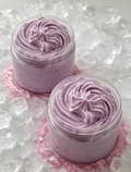 Two small clear jars filled with lavender-pink whipped cream or body butter, swirled on top of pink glitter and surrounded by ice cubes.