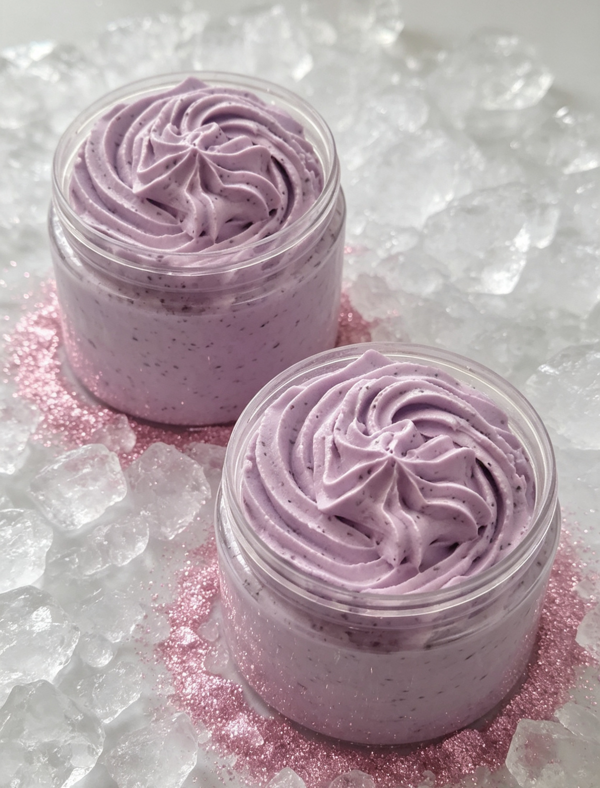 Two small clear jars filled with lavender-pink whipped cream or body butter, swirled on top of pink glitter and surrounded by ice cubes.