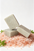 Three rectangular bars of speckled light beige soap stacked on a bed of pink Himalayan salt crystals with small green herb sprigs, photographed on a clean white background.