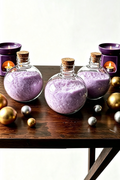 Three round plastic bottles filled with lavender bath salts, sealed with cork stoppers, arranged on a dark wooden table with candles and metallic ornaments in the background.