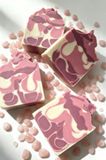 Four rectangular bars of marbled pink and cream soap arranged on a white surface with scattered pink stones. The soap has a swirled pattern in shades of dusty pink, mauve, and warm off-white.