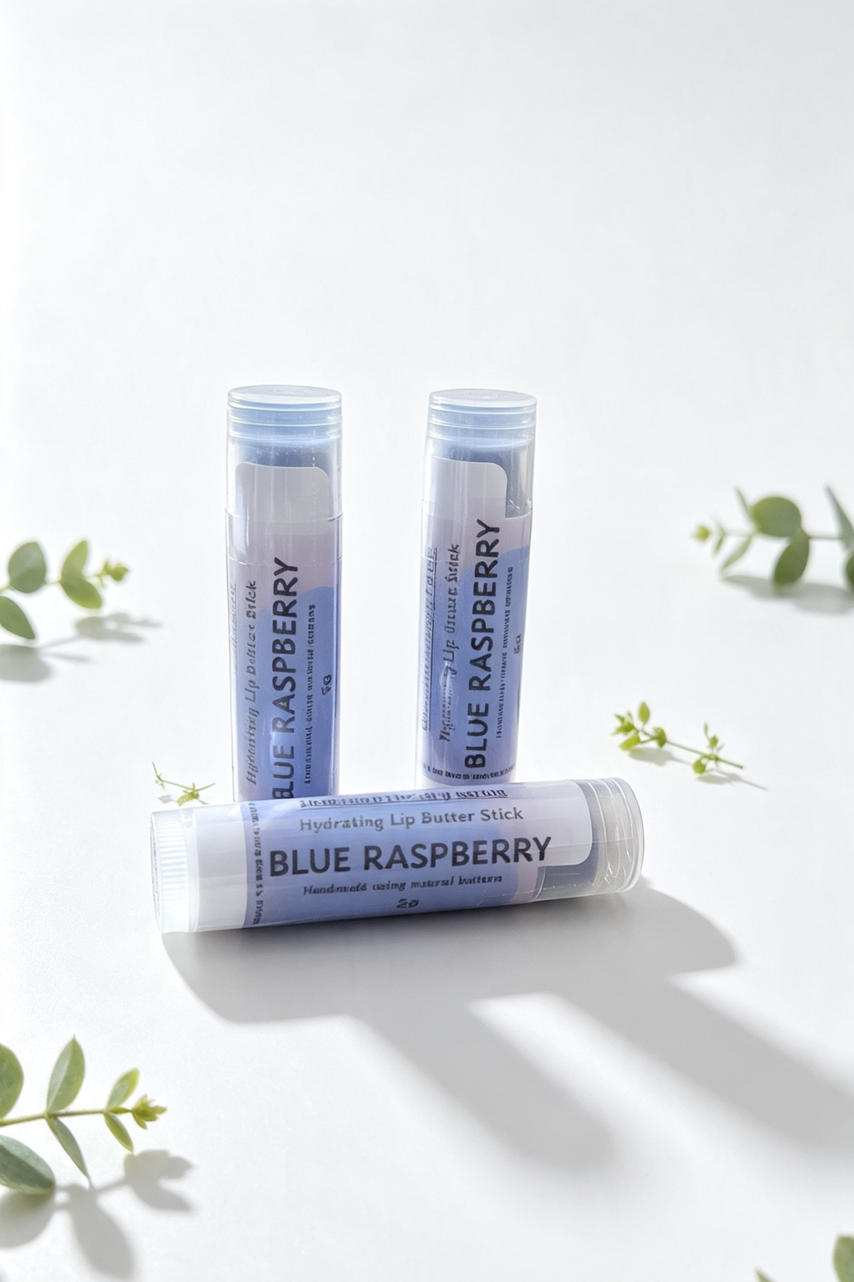3 lip balm sticks on a white background surrounded by sprigs of green leaves