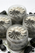 Four clear plastic jars filled with a swirled gray and off-white whipped dessert, arranged on a white surface with scattered black charcoal pieces and a few thin green sprigs.