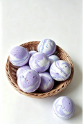Purple and white marbled bath bombs arranged in a round woven wicker basket on a clean white background, with one bath bomb placed outside the basket.