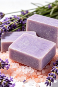Three rectangular lavender soap bars arranged on course pink rock salts with loose lavender sprigs around them on a white background.