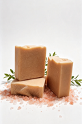Three rectangular bars of natural soap in a warm tan color, arranged on a white surface with scattered pink salt crystals and green leaves.