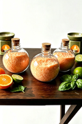 Three round clear plastic bottles with cork stoppers filled with orange bath salts, arranged on a dark wooden table with citrus slices, limes, basil leaves, and green candles in the background.