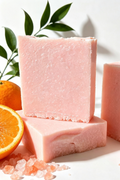 Pinky Orange handmade soap bars stacked on a white surface with a whole orange, a sliced orange, and pink salt crystals nearby, with green leaves in the background.