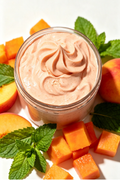 A jar of peach-colored whipped body butter or cream, styled with fresh peach slices, diced mango, and mint leaves on a clean white background.