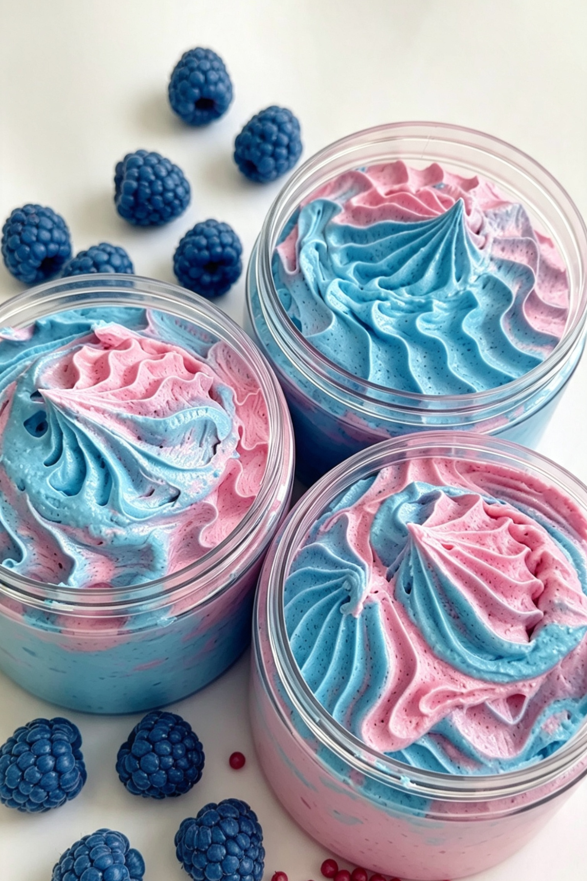 Three clear jars filled with whipped pink and blue cream, arranged on a white surface with scattered Blue Raspberries and Pink Peppercorns around them.