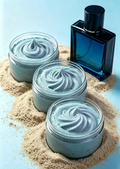 Three open jars filled with whipped pale blue cream are arranged in sand beside a dark blue rectangular perfume bottle on a light blue surface.
