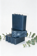 Three stacked rectangular bars of dark blue soap with a textured, wavy top surface, styled with green eucalyptus leaves on a clean white background.