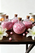 Five round plastic bottles with cork stoppers filled with pink bath salts, arranged on a dark wooden table with white lilies and candles in the background.